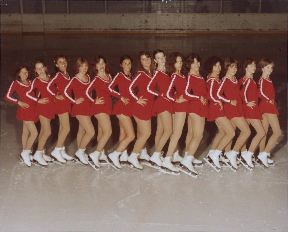 Fifteen Hayden Synchro skaters in red dresses lined up in profile on the ice, 1978 to 1979