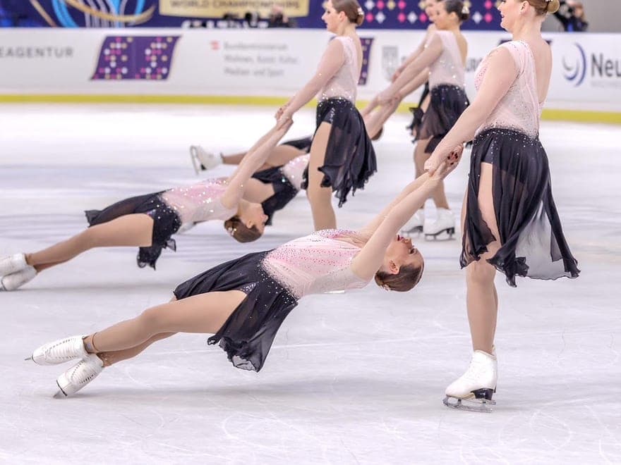 A standing skater holds the hands of a teammate leaning far back, low over the ice, as other pairs do the same behind them